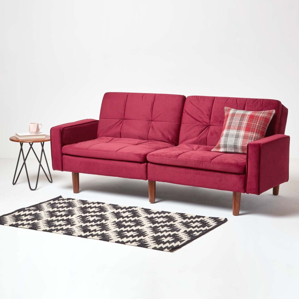 Red murphy sofa bed with a scatter cushion on it, a rug infront of it and a side table. Table has a book and a mug on it.